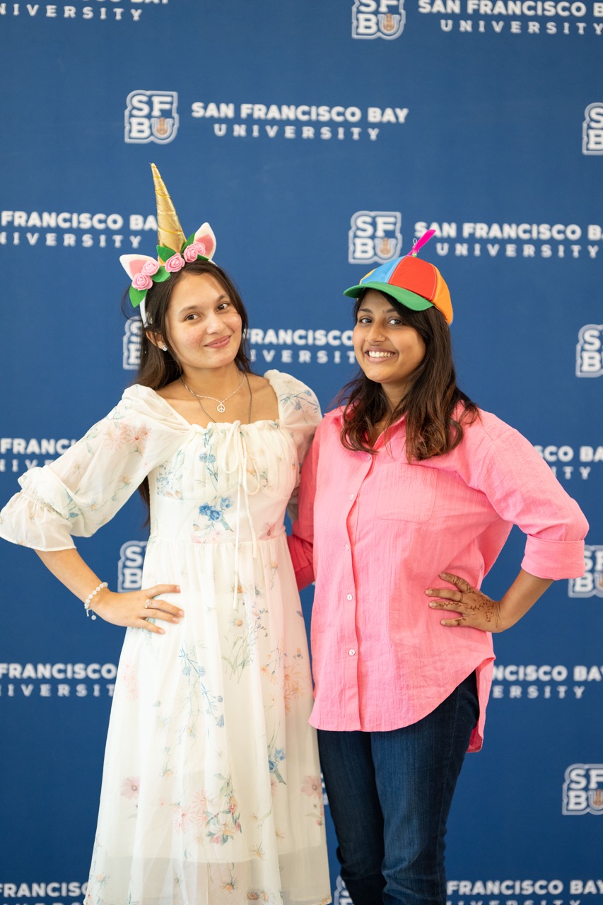 Riddhita Chakma and Priyangka Sarmi wear fun props and pose in front of the SFBU backdrop. 