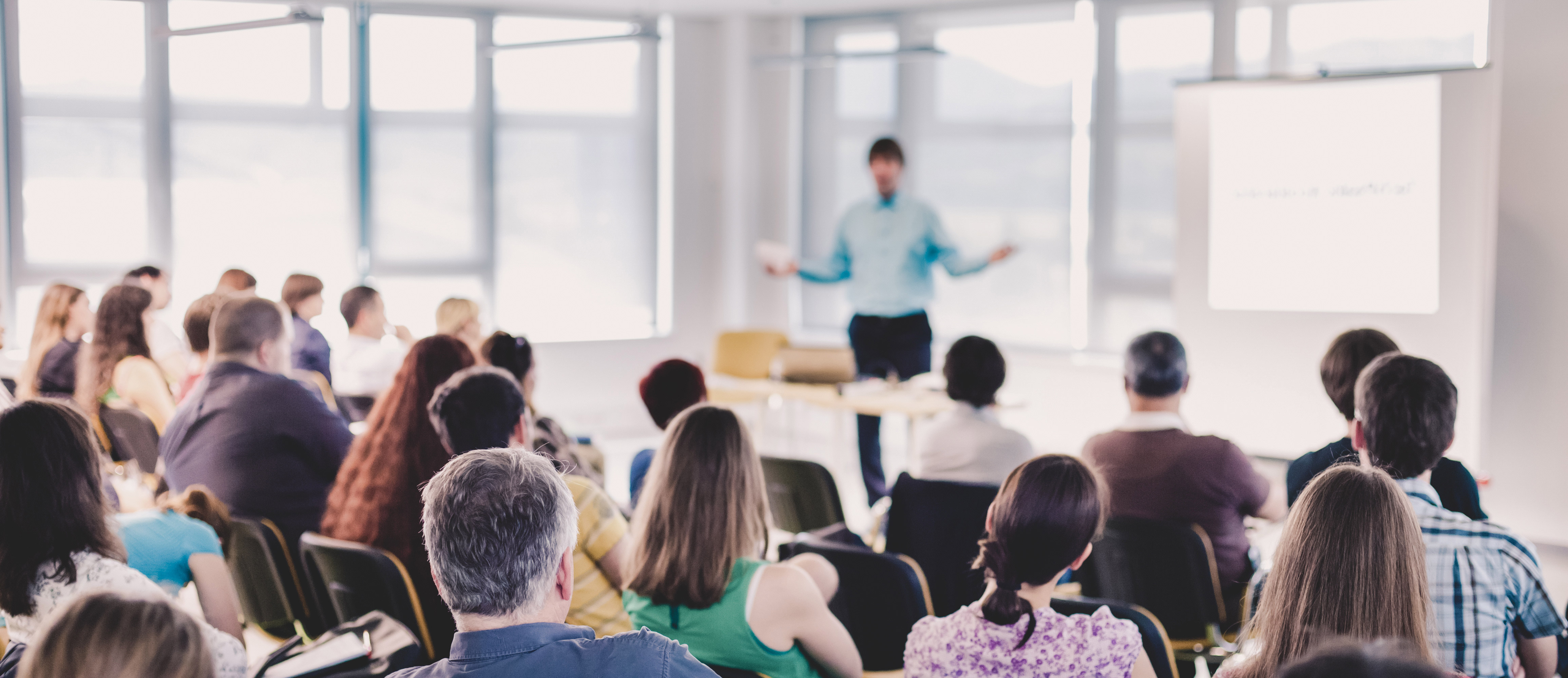 Out of focus presenter in front of crowd with people sitting on chairs.