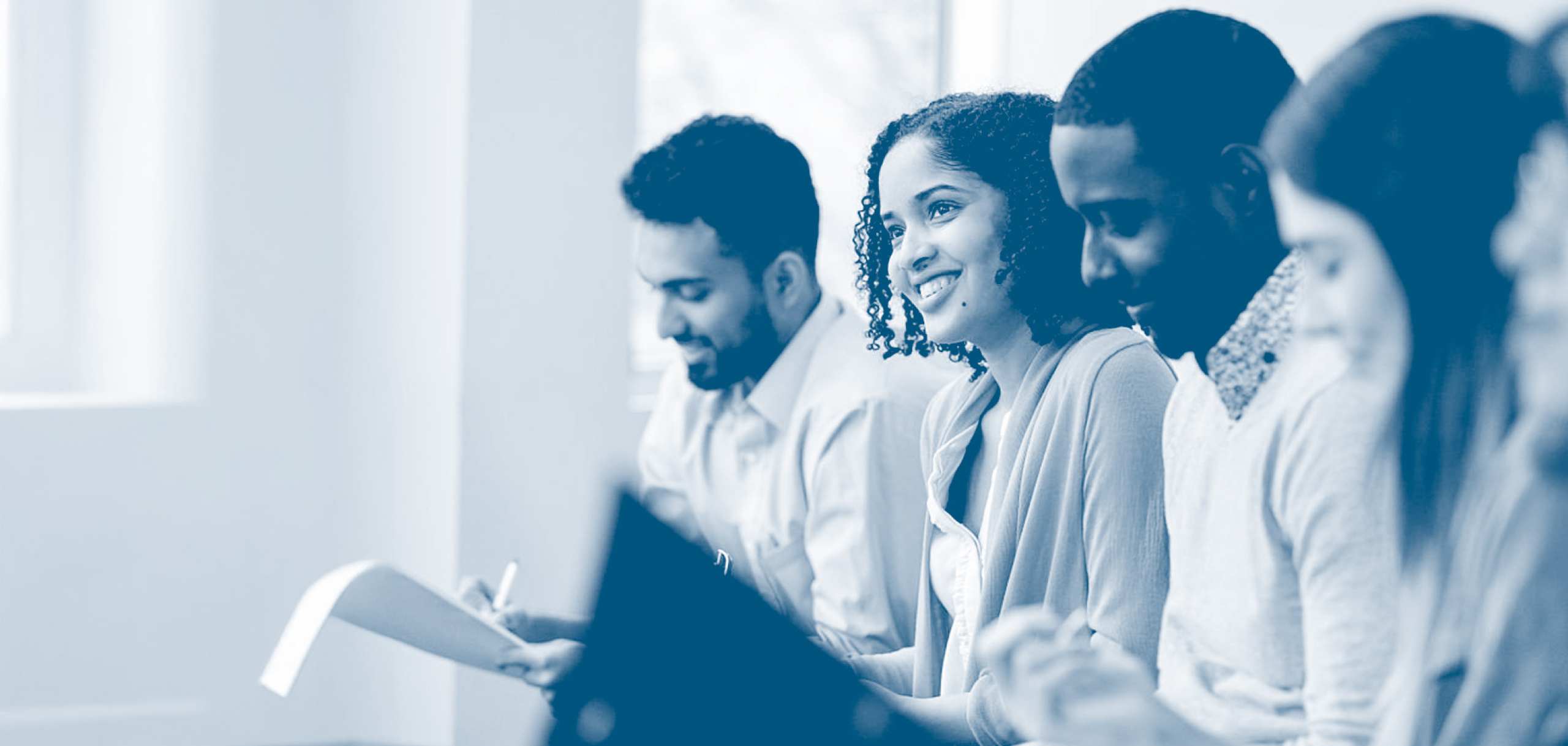 A group of students sit side by side in a classroom, smiling and reviewing notes, with a focus on a student in the center engaged in discussion.
