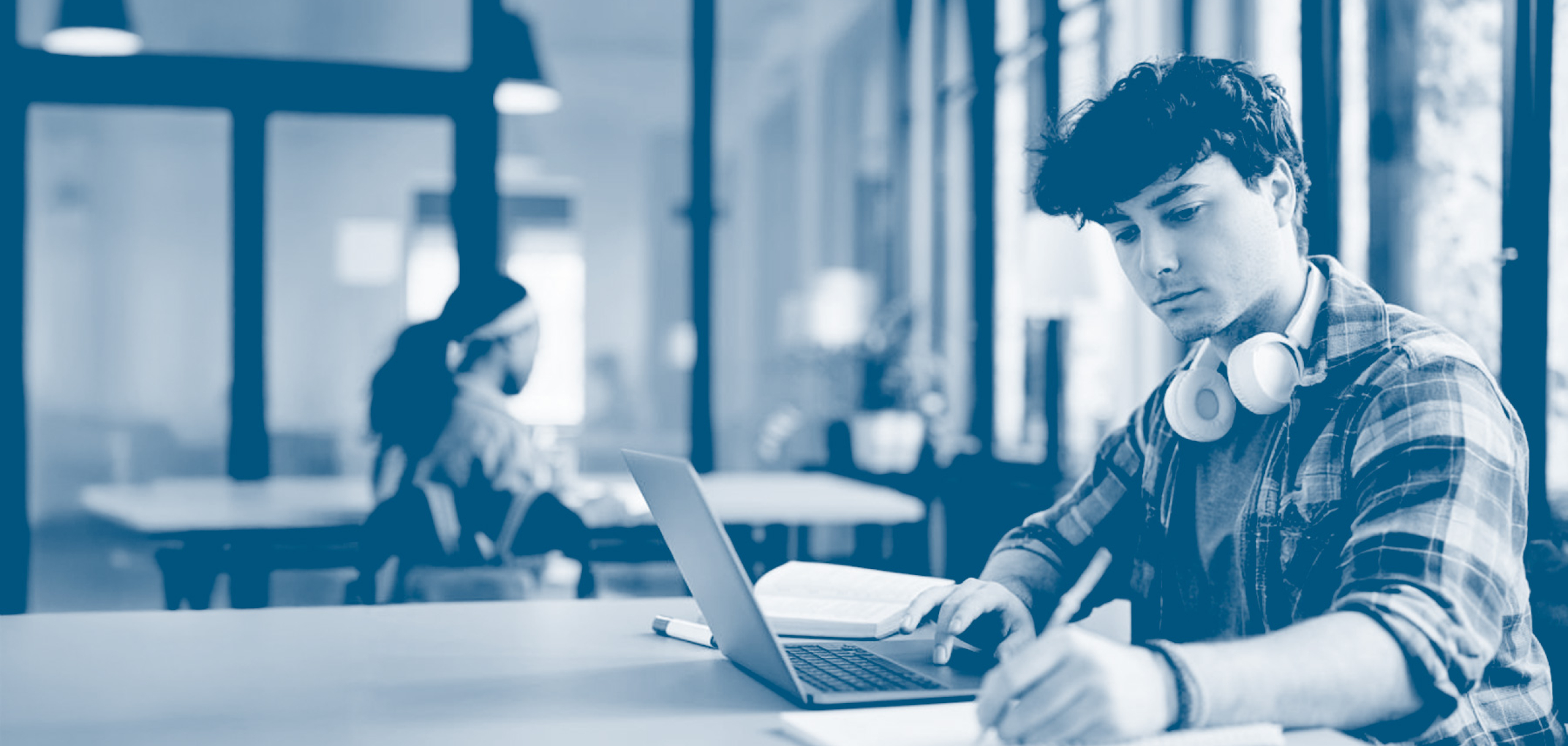 Student wearing headphones studies at a laptop in a modern campus workspace, taking notes while seated at a table.