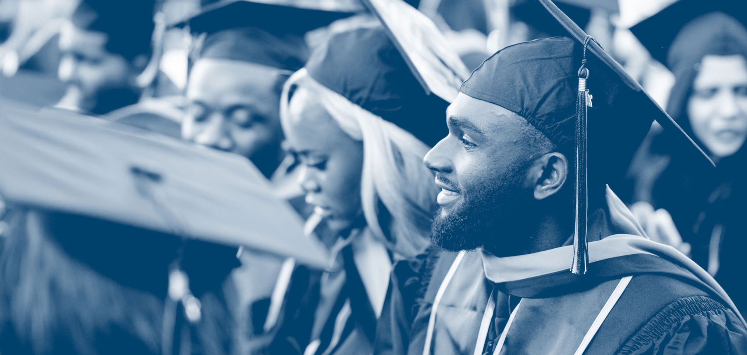 Graduates wearing caps and gowns sit together during a commencement ceremony, smiling and looking ahead, with a focus on a bearded graduate in the foreground.