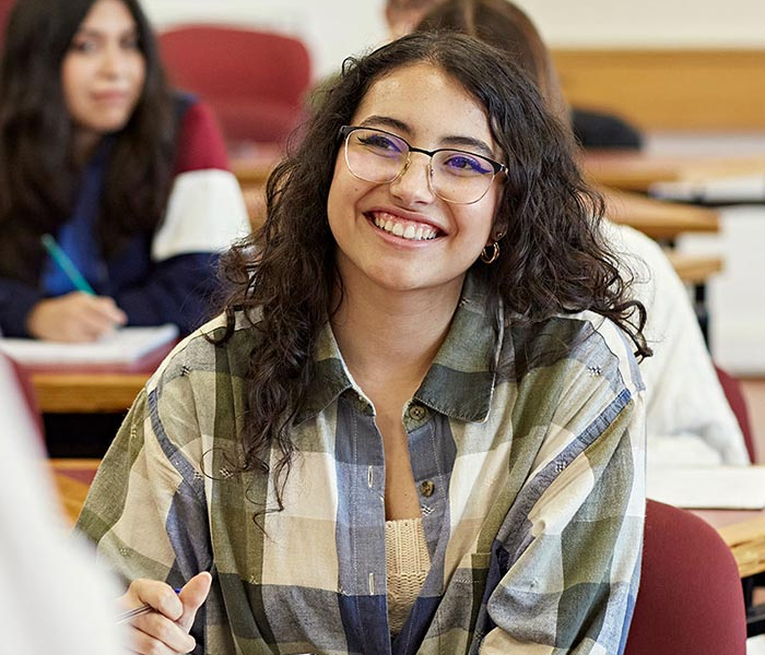 Smiling student with glasses sits in a classroom, holding a pen and looking toward the camera while classmates study in the background.