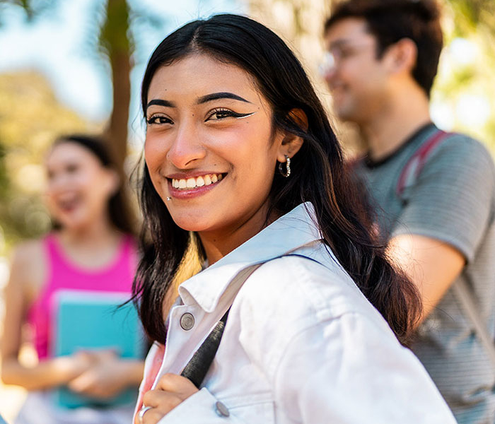 Smiling student outdoors with a backpack looks over her shoulder, with other students blurred in the background on campus.
