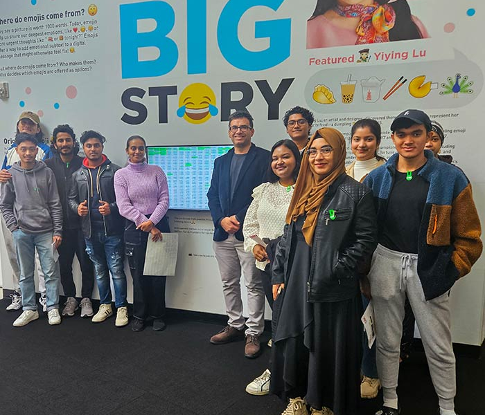 Group of students and an instructor stand together in front of a museum-style display about emojis, posing for a photo.