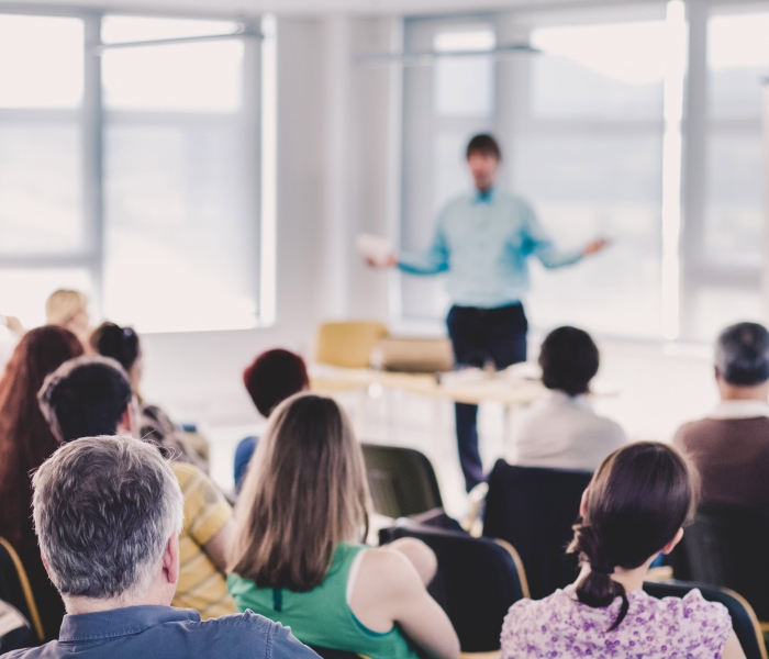 Out of focus presenter in front of a crowd that is sitting on chairs.