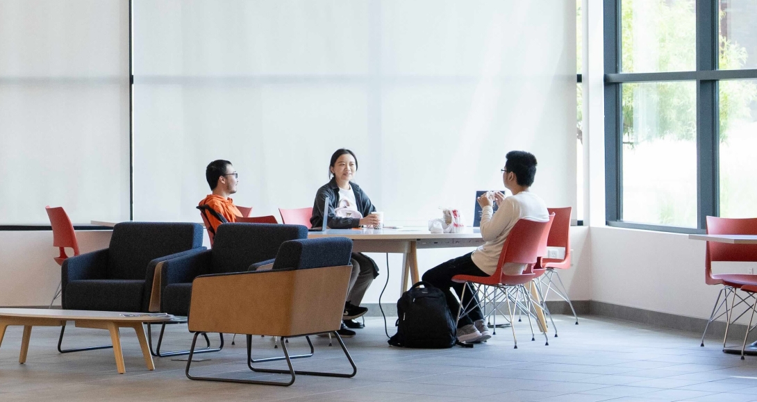 Three students are casually chatting in the lobby of the SFBU campus. The lobby is modern and welcoming, with comfortable seating areas and natural light filtering through large windows. 