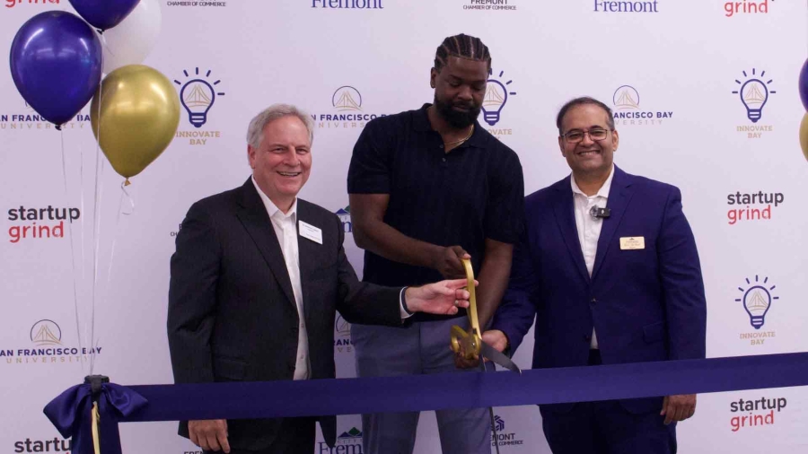 Three men smile during a ribbon-cutting ceremony, with one holding large scissors to cut a blue ribbon, celebrating the opening of an innovation event. Balloons and a branded backdrop with logos for San Francisco Bay University, Startup Grind, and Innovate Bay are visible.